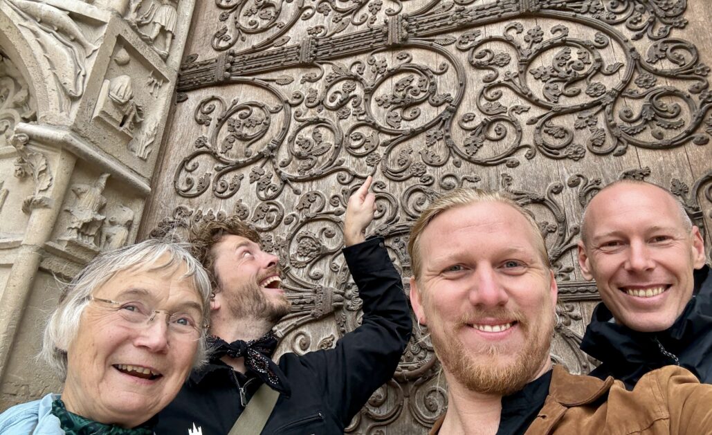 Tre menn og en kvinne står utenfor en gammel kirkedør. De smiler.