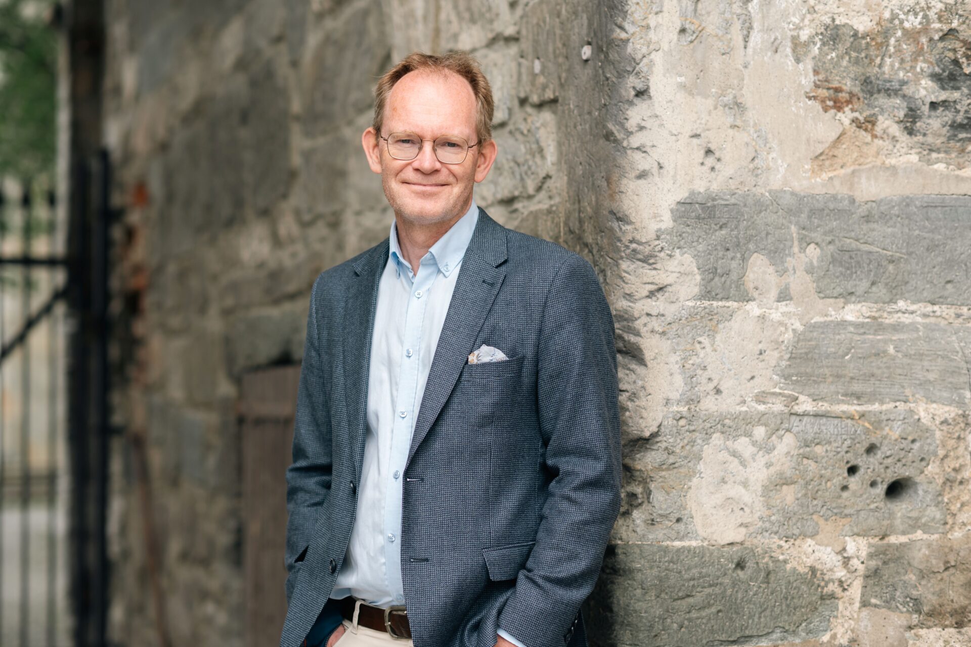 En mann i dressjakke står og smiler foran et gammelt murbygg. En mann i dressjakke står og smiler foran et gammelt murbygg.