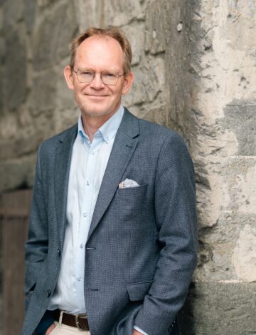 En mann i dressjakke står og smiler foran et gammelt murbygg.