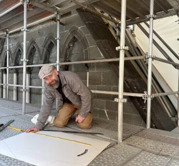 En håmdverker sitter på kne på et stillas og ser på en arbeidstegning.