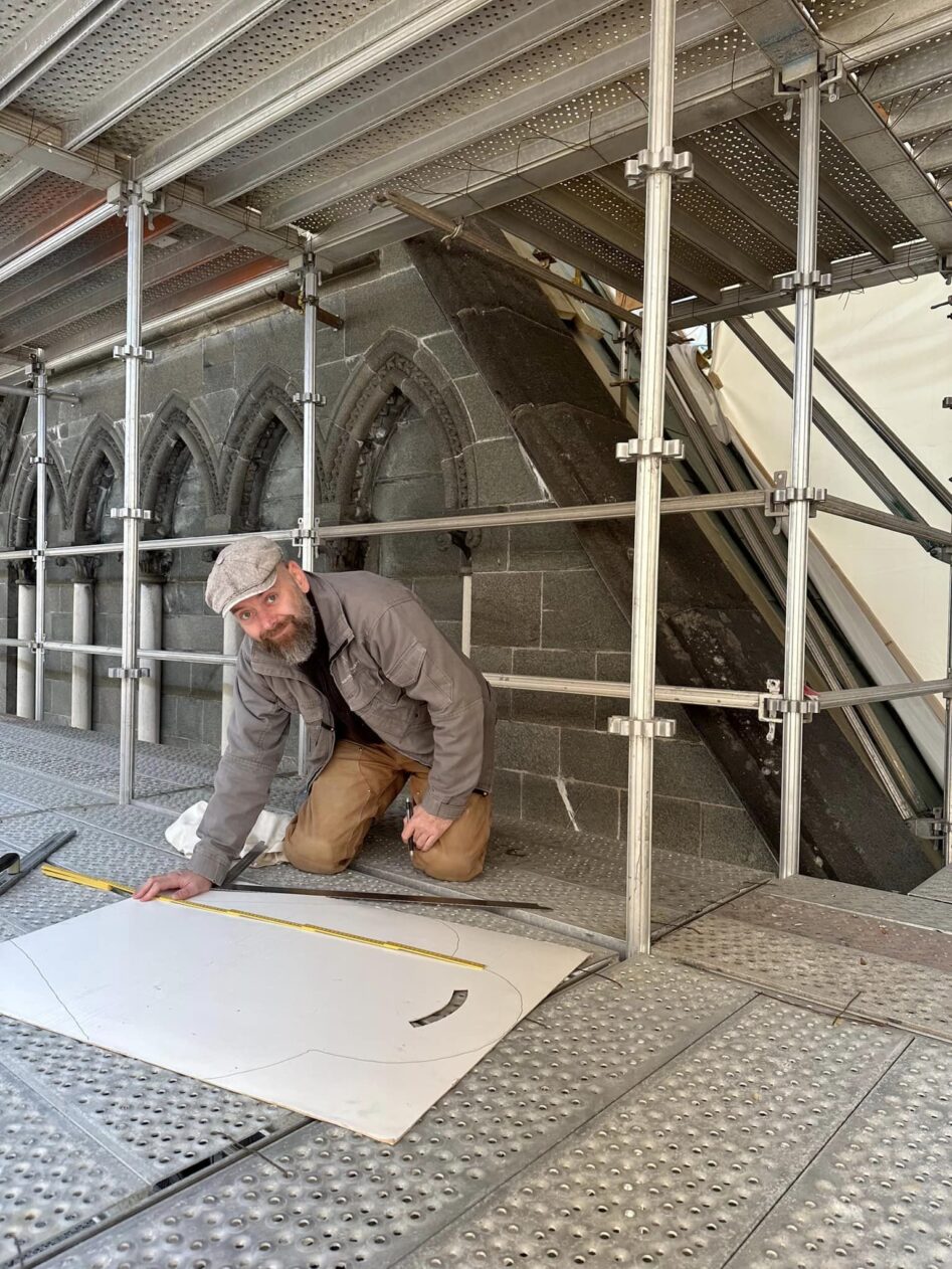 En håmdverker sitter på kne på et stillas og ser på en arbeidstegning.